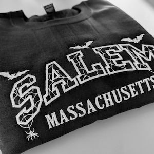 May include: A black crewneck sweatshirt featuring white embroidered text that reads SALEM MASSACHUSETTS. The letters of SALEM are filled with a spiderweb pattern, and the design includes small embroidered bats and spiders hanging from the letters.