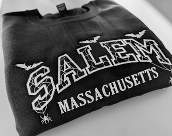 May include: A black crewneck sweatshirt featuring white embroidered text that reads SALEM MASSACHUSETTS. The letters of SALEM are filled with a spiderweb pattern, and the design includes small embroidered bats and spiders hanging from the letters.