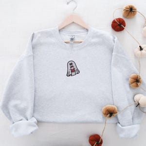 May include: A gray sweatshirt with a small embroidered ghost holding a cup of coffee. The ghost has a surprised expression on its face.
