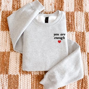 Op de afbeelding: Een lichtgrijs sweatshirt met ronde hals en de zwarte tekst you are enough geborduurd op de linkerborst, geplaatst boven een klein, effen rood hartje. Het kledingstuk heeft lange mouwen en een geribbelde kraag, boorden en zoom.