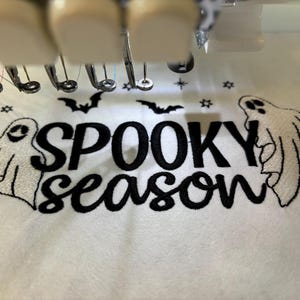 May include: White fabric with black embroidery of the text "Spooky Season" with two white ghosts on either side of the text. There are also small black stars and bats embroidered on the fabric.