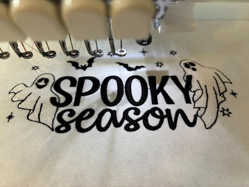 May include: White fabric with black embroidery of the text "Spooky Season" with two white ghosts on either side of the text. There are also small black stars and bats embroidered on the fabric.