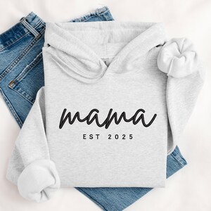 K&ouml;nnte beinhalten: Ein grauer Kapuzenpullover mit dem Text "mama est 2025" in Schwarz.