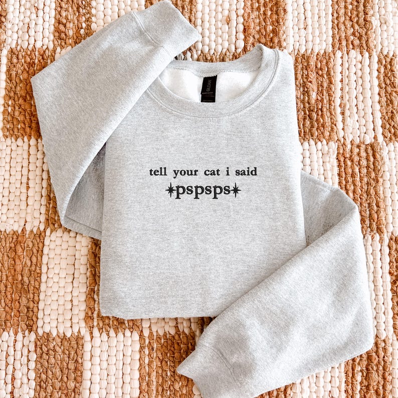 Pu&ograve; includere: Una felpa girocollo grigio chiaro con testo ricamato in nero che recita "tell your cat i said *pspsps*" con piccoli simboli a stella su entrambi i lati della parola pspsps. Il capo presenta maniche lunghe e un colletto a coste.