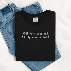Könnte beinhalten: Schwarzes T-Shirt mit weißem Text, der "Will burn sage and *bridges as needed*" lautet, mit einem Stern auf jeder Seite des Wortes "bridges".