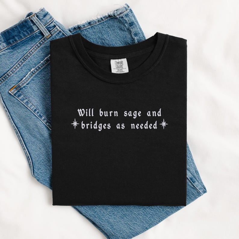 May include: Black t-shirt with white text that reads "Will burn sage and *bridges as needed*" with a star on either side of the word "bridges".