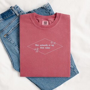 Pode incluir: Uma camiseta vinho com o texto "This actually is my first rodeo" em um losango branco com detalhes florais.
