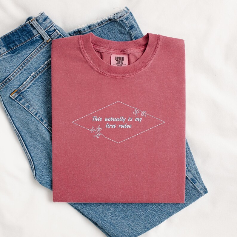 Pode incluir: Uma camiseta vinho com o texto "This actually is my first rodeo" em um losango branco com detalhes florais.