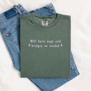 May include: A green t-shirt with the text "Will burn sage and *bridges as needed*" in white.
