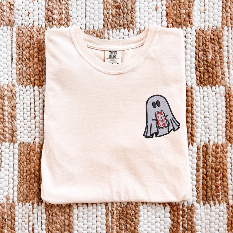 Op de afbeelding: Een licht perzikkleurig t-shirt met een kleine afbeelding op de linkerborst van een grijze geest die een rood Coca-Cola-blikje vasthoudt. Het shirt heeft een ronde hals en een Comfort Colors-merklabel aan de binnenkant van de kraag.