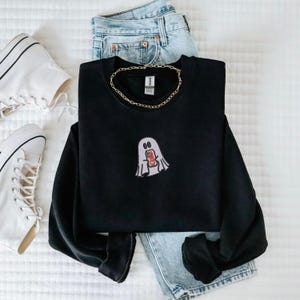 May include: Black sweatshirt with a white ghost holding a red and white can. The sweatshirt is laying on a white surface with a pair of white sneakers and blue jeans.