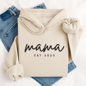 K&ouml;nnte beinhalten: Ein beiger Hoodie mit schwarzem kursivem Text, der "mama est 2025" lautet.