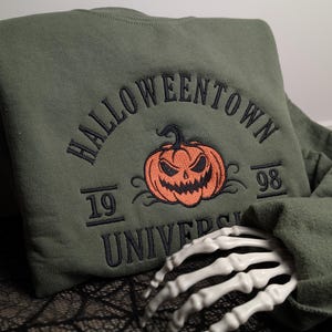 May include: An olive green crewneck sweatshirt featuring dark green embroidered text that reads HALLOWEENTOWN UNIVERSITY with the numbers 19 and 98 on either side. A central orange embroidered jack-o'-lantern pumpkin sits above the text.