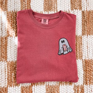 Op de afbeelding: Een rood t-shirt met ronde hals en een kleine geborduurde afbeelding op de linkerborst van een lichtgrijs spookje dat een rood blikje Coca-Cola Classic frisdrank vasthoudt. Het shirt heeft een Comfort Colors merklabel in de nek.