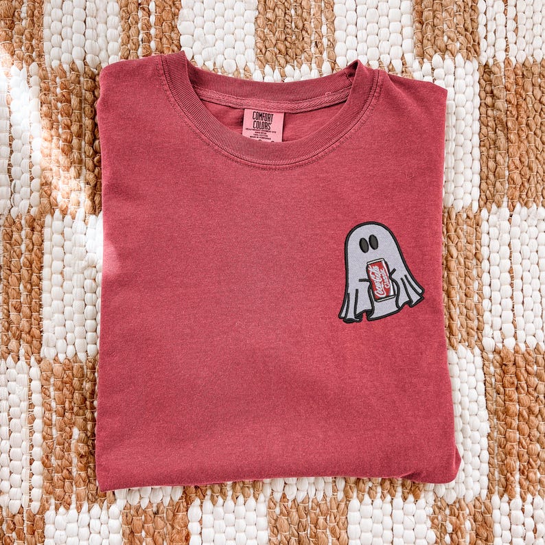 Op de afbeelding: Een rood t-shirt met ronde hals en een kleine geborduurde afbeelding op de linkerborst van een lichtgrijs spookje dat een rood blikje Coca-Cola Classic frisdrank vasthoudt. Het shirt heeft een Comfort Colors merklabel in de nek.