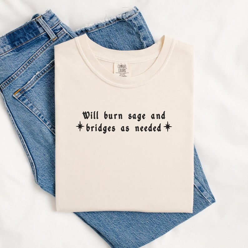 May include: A cream-colored t-shirt with the text "Will burn sage and *bridges as needed*" in black.