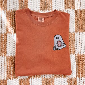 Op de afbeelding: Een roestkleurig t-shirt met korte mouwen en een ronde hals. Op de linkerborst bevindt zich een geborduurde afbeelding van een lichtgrijze geest die een rood frisdrankblikje vasthoudt met de tekst Coca-Cola. Het shirt heeft een Comfort Colors label.