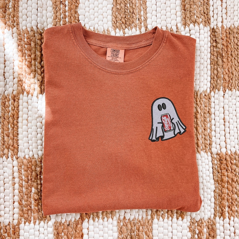 Op de afbeelding: Een roestkleurig t-shirt met korte mouwen en een ronde hals. Op de linkerborst bevindt zich een geborduurde afbeelding van een lichtgrijze geest die een rood frisdrankblikje vasthoudt met de tekst Coca-Cola. Het shirt heeft een Comfort Colors label.