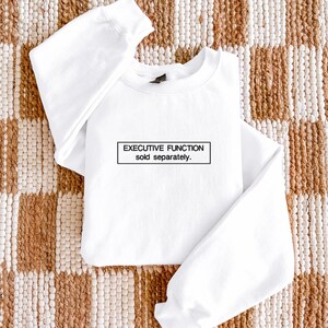 May include: A white long-sleeve crewneck sweatshirt featuring a black rectangular box on the chest. Inside the box, the text reads "EXECUTIVE FUNCTION sold separately." in a clean, sans-serif font. The garment has ribbed cuffs and a ribbed neckline.