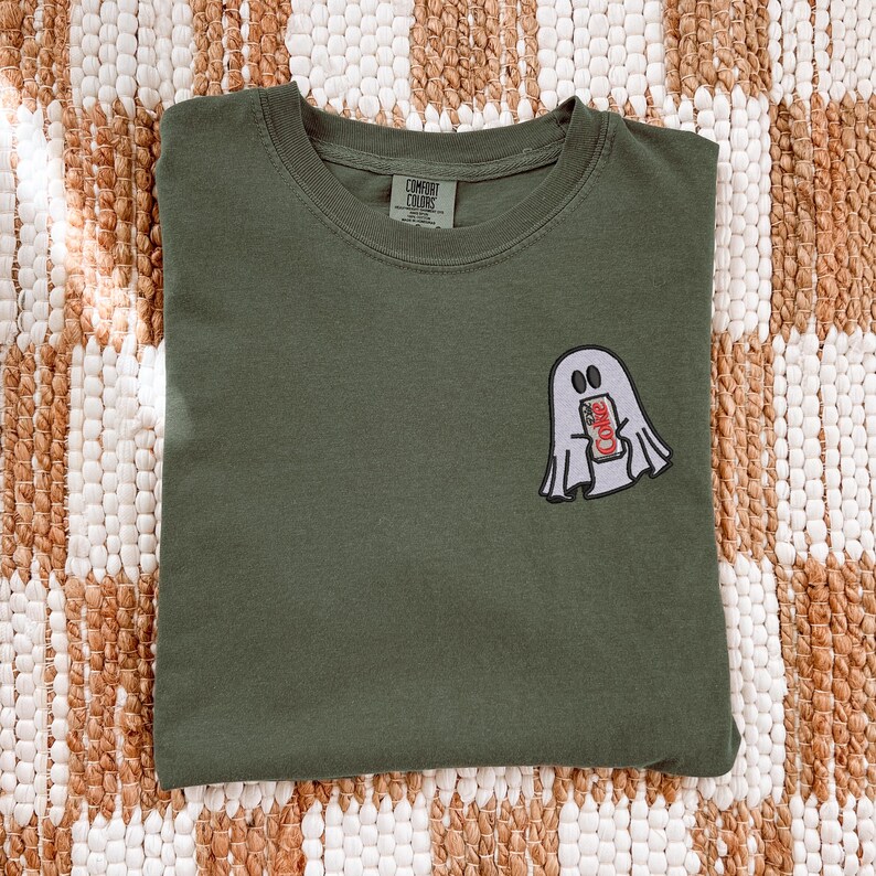 Op de afbeelding: Een olijfgroen t-shirt met ronde hals en een geborduurde afbeelding van een witte geest die een zilveren frisdrankblikje vasthoudt met een rood etiket waarop Diet Coke staat. Het Comfort Colors merklabel is aan de binnenkant van de hals gedrukt.