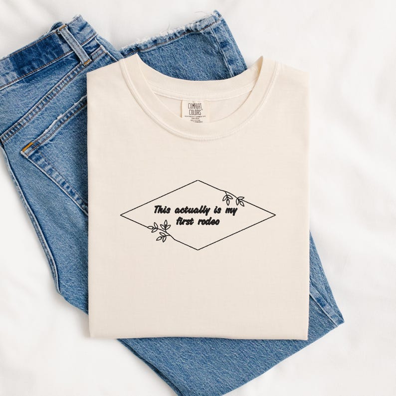 Pode incluir: Uma camiseta cor creme com o texto "This actually is my first rodeo" dentro de um diamante com detalhes florais.