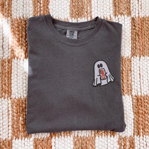 Op de afbeelding: Een antracietgrijs T-shirt met ronde hals en een kleine geborduurde afbeelding op de linkerborst. Het ontwerp toont een wit spook met zwarte omlijning dat een rood-zilveren frisdrankblikje vasthoudt met de tekst Diet Coke erop.