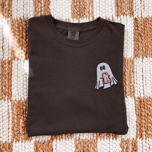 Op de afbeelding: Een donker antracietgrijs t-shirt met ronde hals en een kleine geborduurde afbeelding op de linkerborst. De afbeelding toont een lichtgrijze geest die een rood Coca-Cola Classic frisdrankblikje vasthoudt. Het shirt heeft een Comfort Colors label.