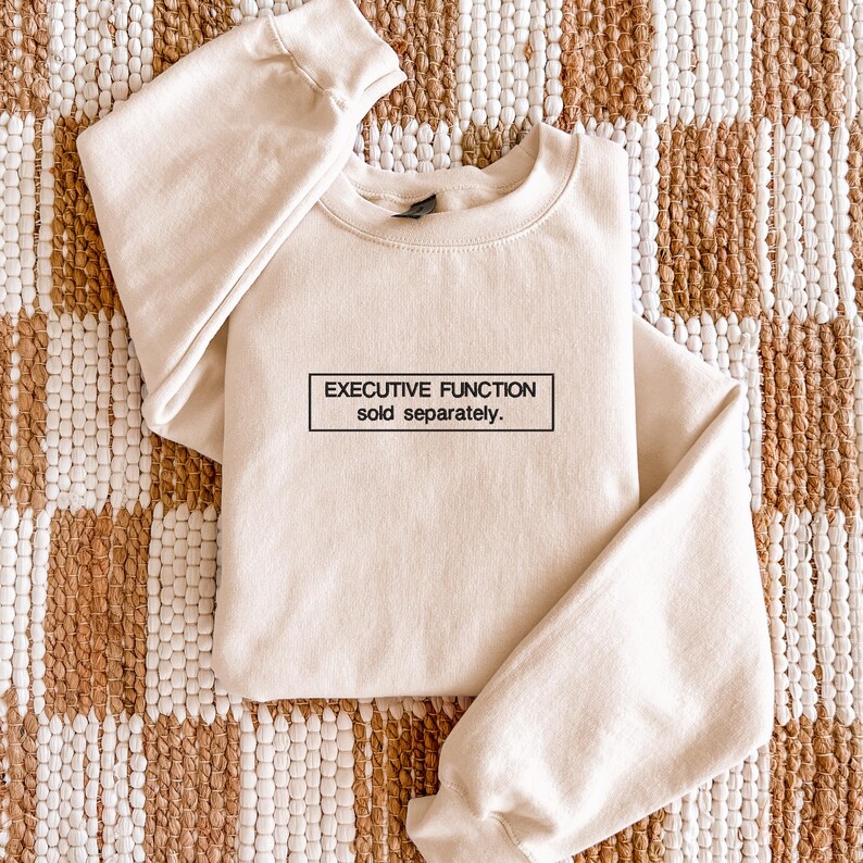 May include: A cream-colored crewneck sweatshirt featuring a black rectangular outline containing the text "EXECUTIVE FUNCTION sold separately." in a clean, sans-serif font. The garment has long sleeves and a ribbed collar, cuffs, and hem.