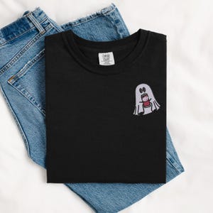 May include: Black t-shirt with a white ghost holding a red can. The ghost has two black eyes and a small mouth. The t-shirt is folded and laid on top of a pair of blue jeans.