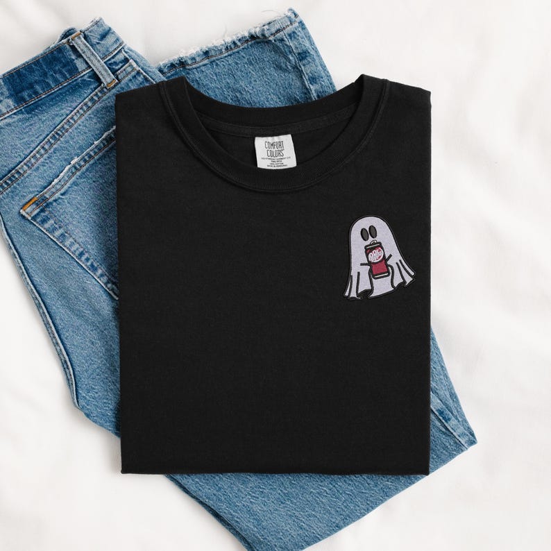 May include: Black t-shirt with a white ghost holding a red can. The ghost has two black eyes and a small mouth. The t-shirt is folded and laid on top of a pair of blue jeans.