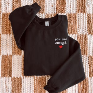 Op de afbeelding: Een zwart sweatshirt met lange mouwen en een ronde hals, voorzien van de witte tekst "you are enough" op de linkerborst, direct boven een klein, effen rood hartje. Het kledingstuk is afgewerkt met een geribbelde kraag en boorden.