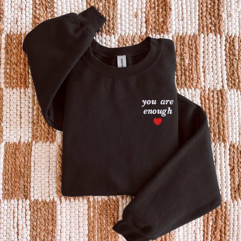 Op de afbeelding: Een zwart sweatshirt met lange mouwen en een ronde hals, voorzien van de witte tekst "you are enough" op de linkerborst, direct boven een klein, effen rood hartje. Het kledingstuk is afgewerkt met een geribbelde kraag en boorden.