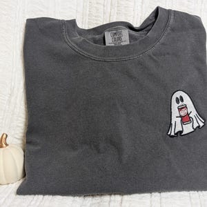 May include: A dark gray t-shirt with a white ghost holding a red and white can. The t-shirt has the text "COMFORT COLORS" on the tag.