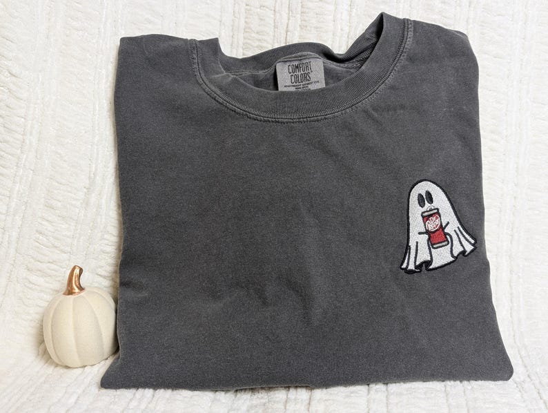 May include: A dark gray t-shirt with a white ghost holding a red and white can. The t-shirt has the text "COMFORT COLORS" on the tag.