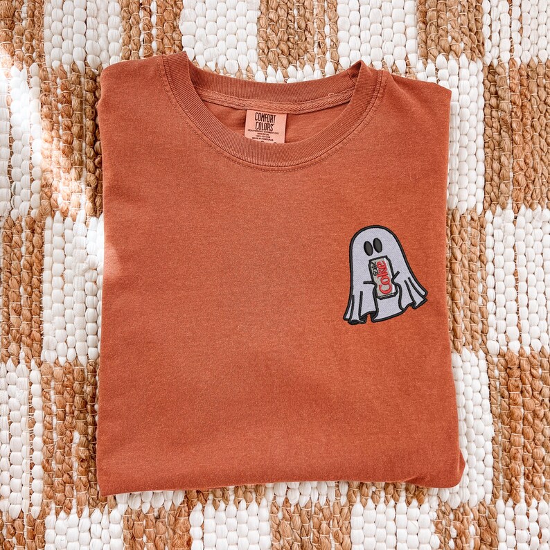 Op de afbeelding: Een roestkleurig T-shirt met ronde hals en een kleine geborduurde afbeelding op de linkerborst van een lichtgrijs spookje dat een rood-wit frisdrankblikje vasthoudt. Het label in de nek bevat de tekst Comfort Colors.