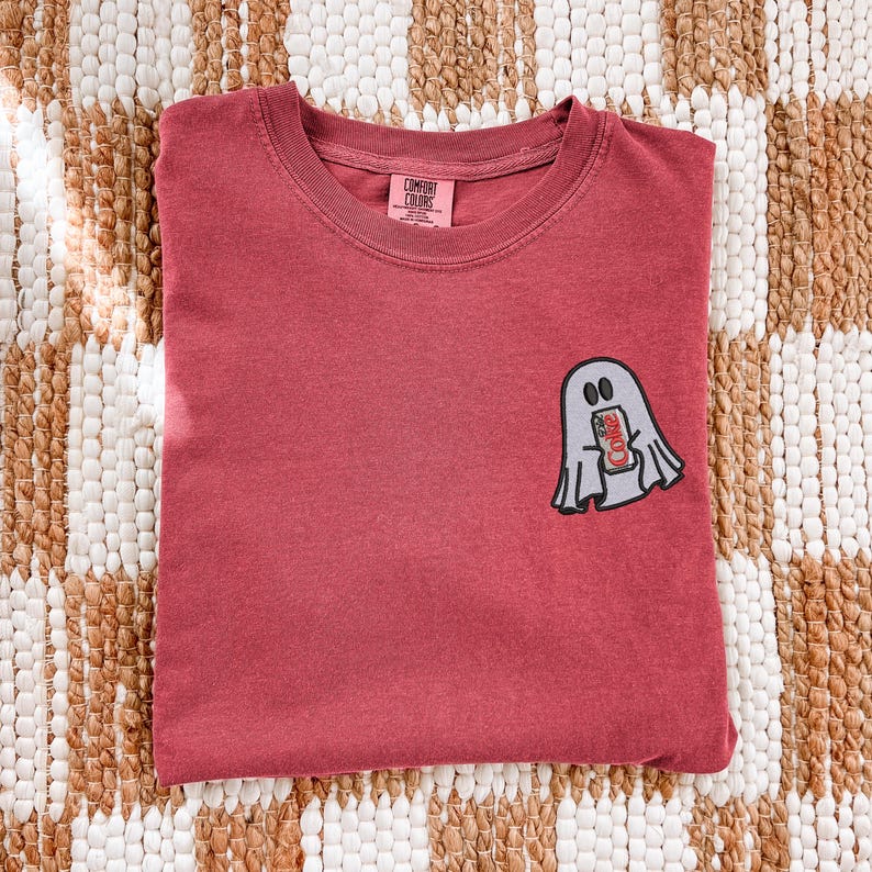 Op de afbeelding: Een baksteenrood T-shirt met ronde hals en een kleine geborduurde afbeelding op de linkerborst van een lichtgrijs spookje dat een rood frisdrankblikje vasthoudt met de tekst Diet Coke. Het Comfort Colors merklabel staat in de nek gedrukt.