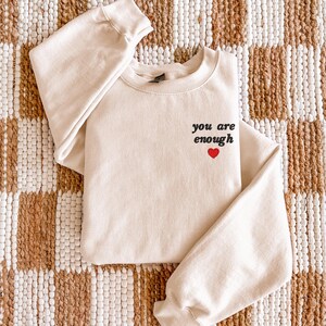 Op de afbeelding: Een cr&egrave;mekleurige sweater met ronde hals voorzien van de zwarte tekst you are enough geborduurd op de linkerborst, vergezeld door een klein, effen rood harticoontje dat direct onder de tekst op de voorzijde is geplaatst.
