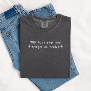 May include: A dark gray t-shirt with the text "Will burn sage and *bridges as needed*" in white.