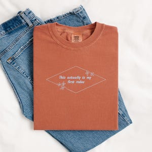 Pode incluir: Uma camiseta cor ferrugem com o texto "This actually is my first rodeo" em um losango branco com detalhes florais.