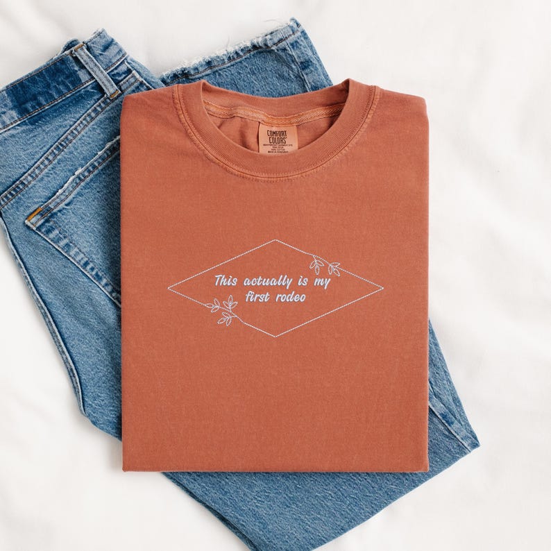 Pode incluir: Uma camiseta cor ferrugem com o texto "This actually is my first rodeo" em um losango branco com detalhes florais.