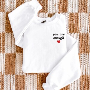 Op de afbeelding: Een wit sweatshirt met lange mouwen en een ronde hals, voorzien van de zwarte geborduurde tekst you are enough op de linkerborst, vergezeld van een klein rood geborduurd hartsymbool direct onder de tekst.