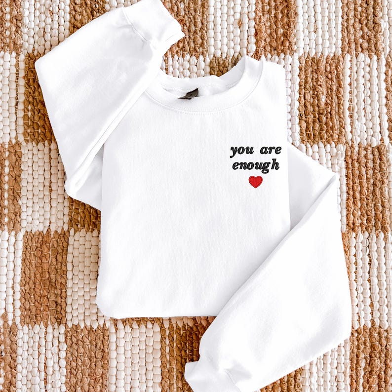 Op de afbeelding: Een wit sweatshirt met lange mouwen en een ronde hals, voorzien van de zwarte geborduurde tekst you are enough op de linkerborst, vergezeld van een klein rood geborduurd hartsymbool direct onder de tekst.