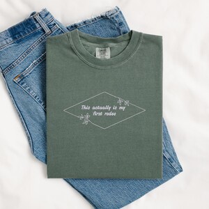 Pode incluir: Uma camiseta verde com o texto "This actually is my first rodeo" em um losango branco com pequenos desenhos florais de cada lado.