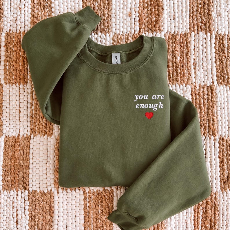 Op de afbeelding: Een olijfgroene sweater met ronde hals en de in wit geborduurde tekst you are enough op de linkerborst, vergezeld van een klein rood hartje direct onder de tekst. Het kledingstuk heeft lange mouwen en een geribbelde kraag.