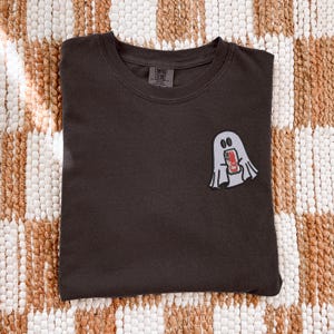 Op de afbeelding: Een donker antracietgrijs t-shirt met ronde hals en een kleine geborduurde afbeelding op de linkerborst. De afbeelding toont een lichtgrijs spookje dat een rood-wit frisdrankblikje vasthoudt met de tekst Coke op de voorzijde van het blikje.