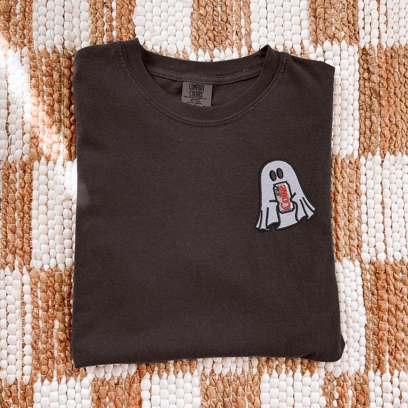 Op de afbeelding: Een donker antracietgrijs t-shirt met ronde hals en een kleine geborduurde afbeelding op de linkerborst. De afbeelding toont een lichtgrijs spookje dat een rood-wit frisdrankblikje vasthoudt met de tekst Coke op de voorzijde van het blikje.