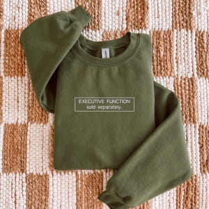 May include: An olive green crewneck sweatshirt featuring a white rectangular outline containing the text "EXECUTIVE FUNCTION" on the top line and "sold separately." on the bottom line, printed in a clean, sans-serif white font.