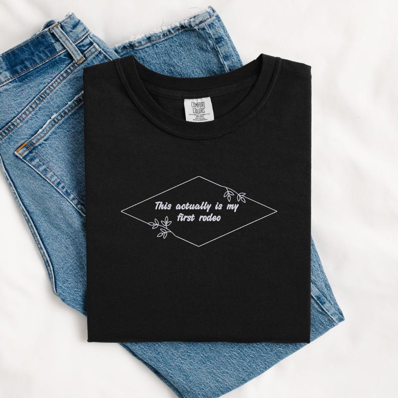Pode incluir: Camiseta preta com texto branco que diz "This actually is my first rodeo" dentro de um diamante com detalhes florais.
