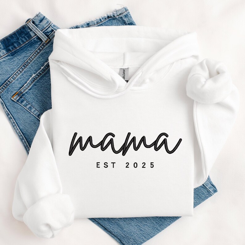 K&ouml;nnte beinhalten: Wei&szlig;er Kapuzenpullover mit dem Text "mama est 2025" in Schwarz.