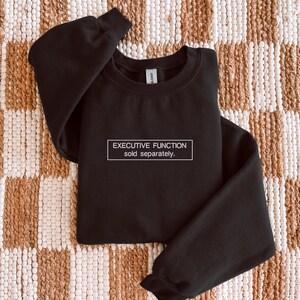 May include: A black long-sleeve crewneck sweatshirt featuring a white rectangular outline on the chest. Inside the rectangle, the text reads "EXECUTIVE FUNCTION" on the top line and "sold separately." on the bottom line in a white sans-serif font.